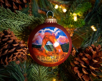 Puede incluir: Una bola de Navidad roja con una imagen de Delicate Arch en el Parque Nacional Arches, Utah. La bola tiene las palabras "Arches National Park" impresas en ella.