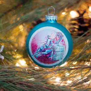 Pode incluir: Um enfeite de Natal azul turquesa com uma borda branca e uma ilustração rosa e azul de um cavalo e um cavaleiro saltando sobre um barril.