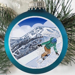 Puede incluir: Adorno navideño azul turquesa con una ilustración blanca y azul de un snowboarder en una ladera de montaña. El adorno tiene un acabado brillante.