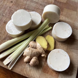 Può includere: Cinque saponette bianche con una texture leggermente macchiata di marrone, disposte su una superficie di legno con un mazzo di steli di citronella e un pezzo di radice di zenzero.