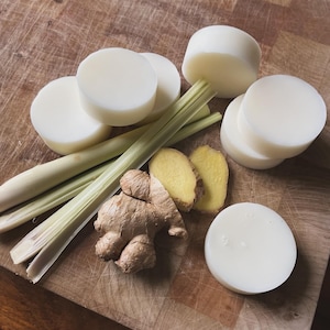 May include: A wooden chopping board with six white round bars of soap, a piece of ginger, and several stalks of lemongrass.