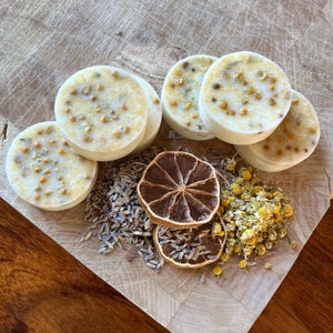 May include: Five round white bath bombs with yellow flecks, a dried lemon slice, and dried chamomile flowers on a wooden surface.