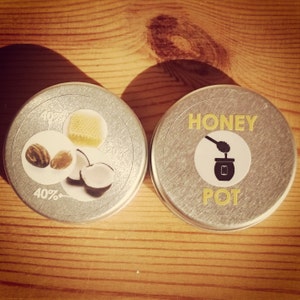 May include: Two silver round tins on a wooden surface. One tin has a graphic of nuts, honeycomb, and coconut with "40%" text. The other tin has "HONEY POT" text and a graphic of a honey dipper and jar.