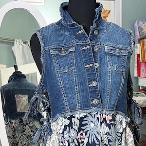 May include: A sleeveless blue denim vest with a frayed hem and metal buttons layered over a long, tiered dress featuring a patchwork of floral patterns in shades of navy blue, white, pink, and muted gold, with side ties on the denim vest.
