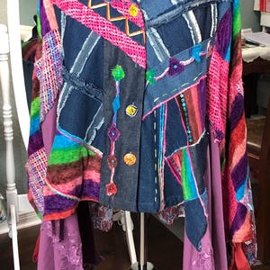 May include: A patchwork denim jacket with a unique, artistic design. The jacket features a mix of denim squares, colorful fabric accents, and decorative stitching. It has a collared neckline, button closure, and a flowing, asymmetrical hemline with lace trim.