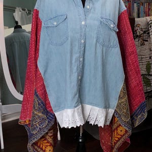 May include: A light blue denim shirt with red and patterned sleeves. The shirt has two front pockets, white buttons, and a white, scalloped hemline. The sleeves are made from a patchwork fabric with red, blue, and yellow designs.