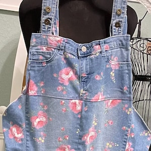 May include: Blue denim overalls with a floral print featuring pink roses. The overalls have adjustable straps with metal buckles.