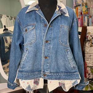 May include: A blue denim jacket with a patchwork collar and hem. The jacket has two chest pockets and brown buttons. The patchwork features floral and geometric patterns in various colors, adding a unique, handcrafted touch to the garment.