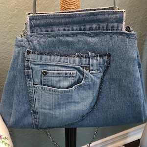 May include: A denim handbag with a silver metal handle and chain strap. The bag is made from recycled blue jeans, featuring a front pocket and visible stitching details. The bag's design showcases the texture and color variations of the denim material.