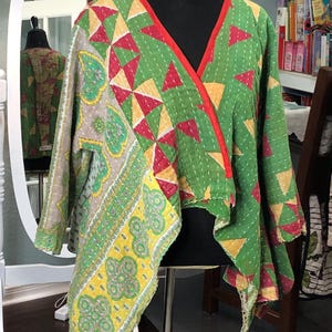 May include: A long-sleeve wrap jacket featuring a patchwork design with green, yellow, and red geometric and paisley patterns. The garment has a V-neckline with a solid red trim and visible white running stitch details throughout the fabric.