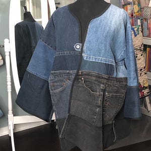 May include: A patchwork denim jacket in various shades of blue, with a black trim and a single button closure. The jacket features pockets from repurposed jeans, creating a unique, upcycled look. The jacket is displayed on a black mannequin.