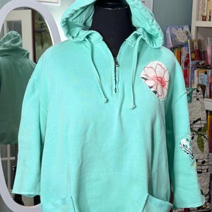 May include: A mint green hooded sweatshirt featuring a quarter-zip neckline with a silver zipper pull, a drawstring hood, and two front pockets. The garment includes a circular floral patch on the left chest and a smaller floral patch on the sleeve.
