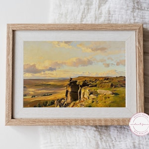 May include: A rectangular landscape painting in a light wood frame depicting a rocky cliff edge overlooking a vast valley under a golden sunset sky. The artwork features warm yellow and orange light hitting the clouds and terrain, with a small figure perched on a rock formation. A circular logo in the bottom right corner contains the text ardent fancy made with love.