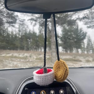Puede incluir: Un amuleto de coche de ganchillo con una rosquilla roja y blanca y una rosquilla amarilla y blanca. El amuleto está colgado del espejo retrovisor de un coche.