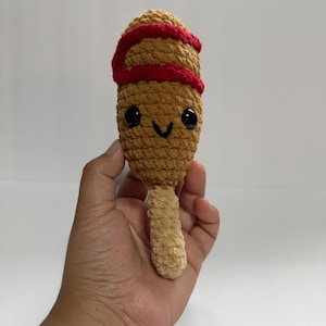Puede incluir: Un juguete de peluche de hot dog de maíz hecho a mano con una cara sonriente, ojos negros y un diseño en espiral rojo. El juguete está hecho de hilo marrón y beige y tiene un palo en la parte inferior.