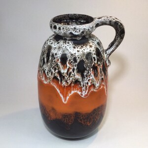 May include: A ceramic pitcher with a handle, featuring a striking color scheme. The top is speckled with black and white, transitioning to orange and then dark brown at the base. The pitcher has a unique, textured glaze.