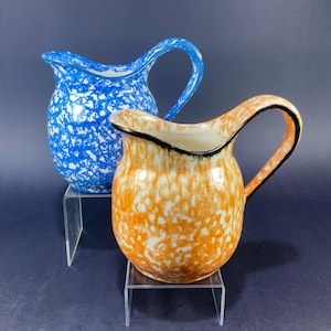 May include: Two ceramic pitchers with speckled designs. One is blue with white speckles, the other is orange with white speckles. Both have curved handles and a black trim around the rim. They are displayed on clear acrylic stands.