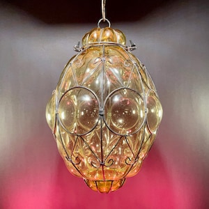 May include: A hanging, ornate, amber-colored glass lantern with a metal filigree cage. The lantern has a rounded, bulbous shape with circular glass accents and a decorative top. The metal cage features intricate scrollwork and geometric patterns.