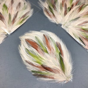 May include: Close-up of several feather bundles. Each bundle features a fan-like arrangement of white, brown, and green feathers. The feathers are arranged in a layered pattern, creating a textured and visually appealing design. The bundles are on a gray surface.