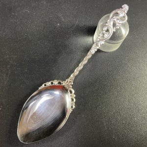 May include: A silver spoon with an ornate, scrolled handle and a twisted stem. The spoon's bowl has a decorative edge. The spoon is resting on a dark surface.