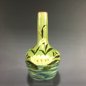 May include: A green ceramic vase with a long neck and a bulbous body. The vase is decorated with a water lily design, featuring white flowers, green lily pads, and blue water. The rim is a reddish-brown color.