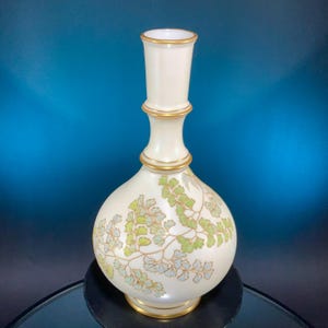 May include: A cream-colored ceramic vase with a bulbous base and a slender neck. The vase is adorned with a gold rim and delicate green and blue-grey fern-like patterns. The vase is approximately 10 inches tall.