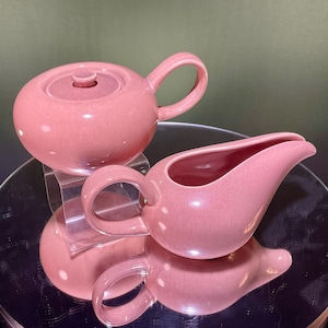 May include: A dusty rose ceramic teapot with a matching lid and a curved handle, paired with a matching dusty rose ceramic creamer pitcher featuring a wide pouring spout and a side handle, both items displaying a smooth, glossy, speckled finish.
