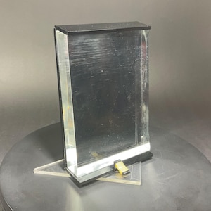 May include: A clear acrylic display case with a black top and base. The case is rectangular and stands upright, supported by a clear acrylic triangle. A small yellow and black component is attached to the base.