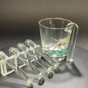 May include: A clear glass mug with a gold rim and a handle, featuring a Dalmatian dog illustration. Beside it are five clear glass stir sticks, each with a decorative white stripe and a black base. The set is displayed on a dark surface.