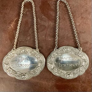 May include: Two oval silver-toned metal bottle tags with attached chains. Each tag features an embossed floral border and engraved text. One tag reads Dry Vermouth, and the other tag reads Sweet Vermouth in a decorative gothic-style font.