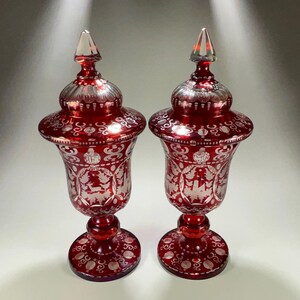May include: Two ornate, ruby red glass urns with lids. The urns feature intricate etched designs and sit on round bases. Each urn has a clear, pointed finial on top. The glass has a reflective quality, enhancing the details.
