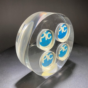 May include: A clear, circular photo display with four circular openings. Each opening contains a blue and white label with the letters "PIC". The display is made of a transparent material, allowing light to pass through.