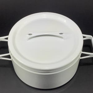 May include: A white enamel cooking pot with a matching lid. The pot has two side handles and a handle on the lid. The pot is round and sits on a black surface. The pot is likely used for cooking.