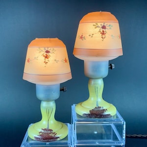 May include: Two vintage table lamps with peach-colored shades adorned with floral patterns. The lamps have a yellow base with a painted tree design and a frosted glass body. The lamps are displayed on clear acrylic stands.