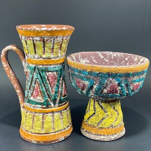 May include: A colorful ceramic pitcher and bowl set. The pitcher has a handle and is decorated with yellow, green, and red geometric patterns. The bowl is supported by a pedestal and features similar colors and designs. The set has a textured, rustic appearance.