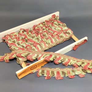 May include: Decorative fringe trim in shades of red, green, and brown, displayed on a white wooden frame. The trim has a textured, looped design and is labeled "VAT DYED". A vintage aesthetic.