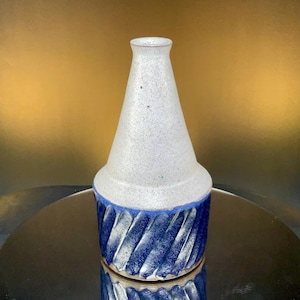 May include: A handmade ceramic vase with a unique design. The upper portion is a speckled white, while the base features a textured, swirling pattern in shades of blue. The vase has a tapered shape and a small opening at the top.