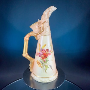 May include: A decorative, cream-colored pitcher with a bamboo-style handle and spout. The pitcher features a floral design with red, pink, and yellow flowers. Gold accents are present on the handle and rim. The pitcher is likely made of ceramic or porcelain.