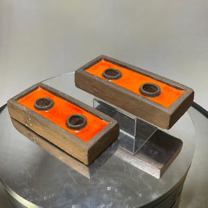 May include: Two rectangular ceramic candle holders with a matte brown exterior and a vibrant orange glazed interior. Each holder features two circular openings for candles, with the dark brown clay rims visible around each opening.