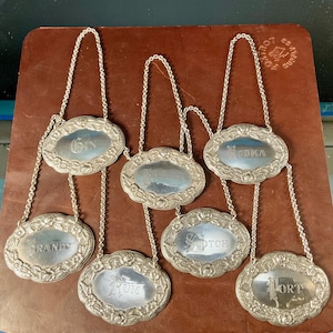 May include: A set of six oval silver-toned metal decanter labels, each suspended by a matching chain. The labels feature embossed floral borders and are engraved with the words GIN, BRANDY, RUM, WHISKEY, SCOTCH, and PORT in a decorative font.