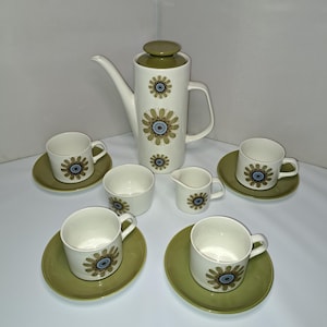May include: A vintage white ceramic coffee set with a floral design. The set includes a tall coffee pot, six cups with saucers, a creamer, and a sugar bowl. The floral pattern features a brown and blue flower design on a white background.