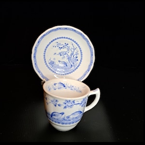 May include: A white teacup and saucer set with blue floral and bird designs. The cup has a handle and the saucer has a scalloped edge. The set is likely vintage and suitable for tea parties or display.