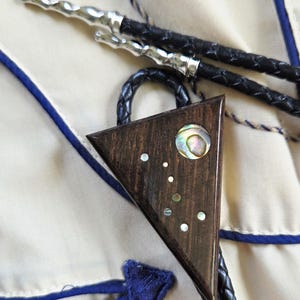 May include: A bolo tie featuring a triangular wooden pendant. The pendant is dark brown with inlaid iridescent shell accents and a large shell circle. The braided black leather cord has silver tips.