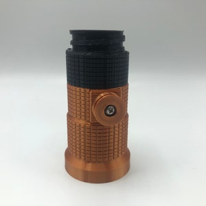 May include: A cylindrical, 3D-printed object with a textured surface. The top section is black, the middle is copper-colored with a grid pattern, and the bottom is copper-colored. A copper-colored circular element with a screw is on the side.