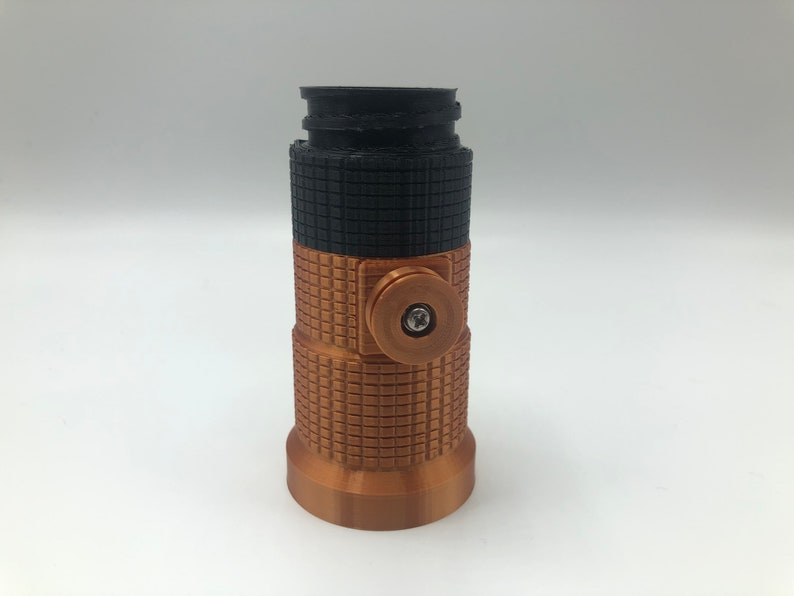 May include: A cylindrical, 3D-printed object with a textured surface. The top section is black, the middle is copper-colored with a grid pattern, and the bottom is copper-colored. A copper-colored circular element with a screw is on the side.
