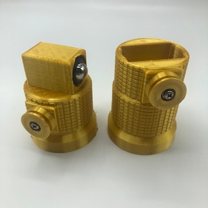 May include: Two gold-colored, cylindrical objects with textured surfaces and circular details. One has a rectangular top with a silver ball, the other has a rectangular opening. Both have a screw detail on the side.
