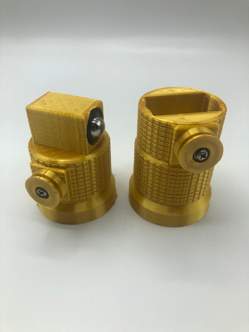 May include: Two gold-colored, cylindrical objects with textured surfaces and circular details. One has a rectangular top with a silver ball, the other has a rectangular opening. Both have a screw detail on the side.