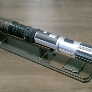 May include: A detailed lightsaber prop with a silver, black, and brown design, resting on a custom metal display stand. The lightsaber features intricate detailing and a metallic finish. The stand is a dark bronze color.