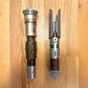 Puede incluir: Dos empuñaduras de sable de luz detalladas, una a la izquierda con detalles en verde oliva y plata, y la otra a la derecha con un envoltorio plateado y marrón similar al cuero. Ambas sobre un fondo de madera.