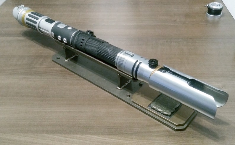 May include: A silver and black lightsaber prop, resting on a custom metal stand. The lightsaber features intricate detailing, including a textured grip and various buttons. The stand is made of dark metal and is mounted on a wooden surface.